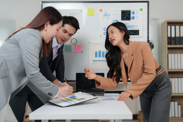 Three asian businesspeople are discussing and working together on a financial report during an office meeting, analyzing charts and graphs displayed on a whiteboard