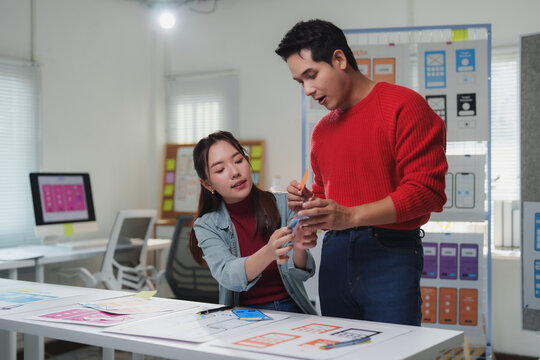 Two asian ux designers are discussing mobile application interface using paper prototypes in office meeting, collaborating on improving user experience and functionality