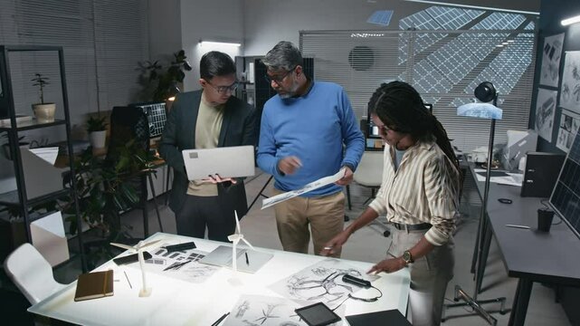 Full shot of diverse team of three engineers using laptop and technical drawings to negotiate ideas about developing sustainable technologies in modern office with multimedia screen and solar panels