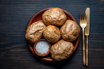 Simple country food, baked potatoes whole in their skins and salt in clay plate on wooden table, closeup, top view