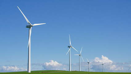 Fototapeta premium Wind turbines spin on rolling hills beneath a clear blue sky with fluffy clouds, symbolizing clean, renewable energy in harmony with nature.