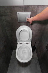 Man is pushing the flush button on a modern, wall-mounted toilet in a bathroom with contemporary grey tiles, highlighting hygiene and sanitation practices. Vertical photo