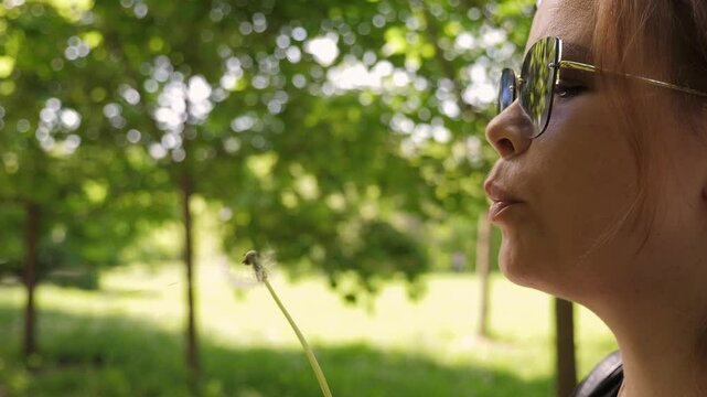 Close-up of a young woman wearing sunglasses blowing off a fluffy dandelion in a sunny park. Fluffy dandelion seeds scatter in different directions.