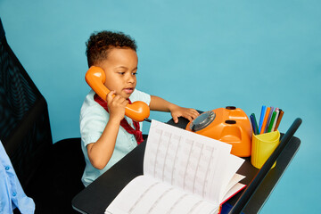 Child holds phone and flips calendar on office desk against blue background showing roleplay and curiosity. Concept of childhood, beauty, creativity, imagination, lifestyle, fun, learning, emotion.