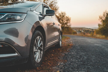 Close-up of modern car parked on roadside with autumn trees in background. Curved country road...