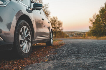 Close-up of modern car parked on roadside with autumn trees in background. Curved country road leads into soft sunset horizon, evoking peaceful travel mood