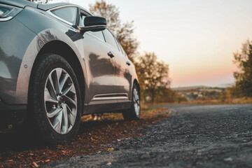Naklejka premium Close-up of modern car parked on roadside with autumn trees in background. Curved country road leads into soft sunset horizon, evoking peaceful travel mood