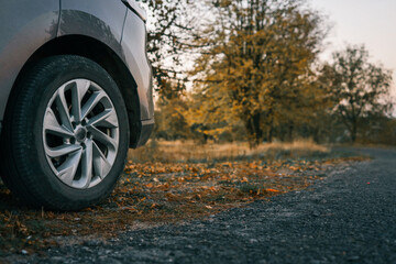 Naklejka premium Close-up of front car wheel on rural road with fallen autumn leaves. Background features soft focus of trees in golden hour light, evoking a calm countryside travel mood