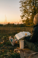 Girl reading book at sunset in autumn field. Bible open on her lap, reusable cup nearby, cozy mood...