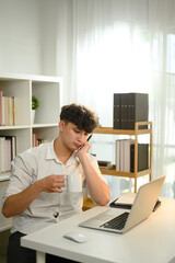 Bored man holding a coffee mug and working with laptop at home office