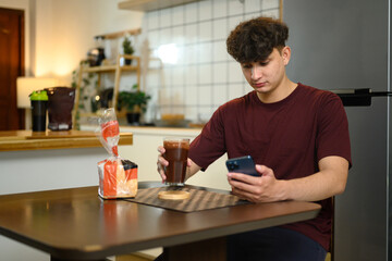 Young man sitting at kitchen table drinking chocolate protein shake and using smartphone