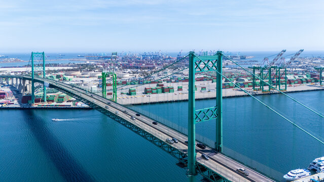 High-angle view of the iconic Vincent Thomas Bridge spanning the Port of Long Beach in California. The turquoise suspension bridge crosses over calm blue water, with stacks of shipping container