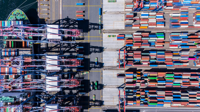 A straight-down aerial photo of shipping cranes operating over a dock filled with colorful cargo containers. The image highlights the symmetry and industrial activity of a modern shipping port.
