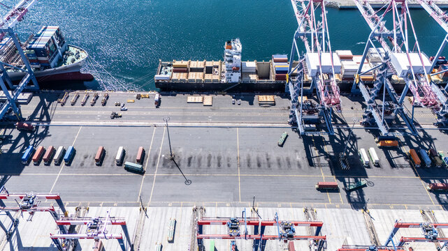 High-angle view of towering shipping cranes moving containers from a large cargo ship docked at a seaport. The deep blue ocean and colorful containers create a dynamic logistics scene.