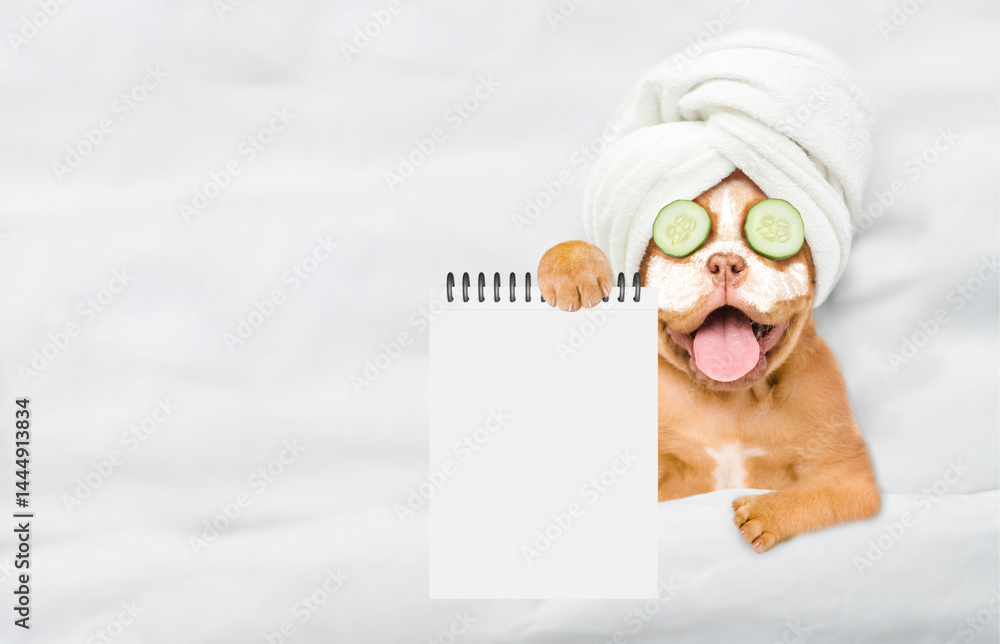 Aufkleber Funny Mastiff puppy with towel on it head and pieces of cucumber on it eyes relaxing on the bed at home and showing empty list. Top down view. Empty space for text
