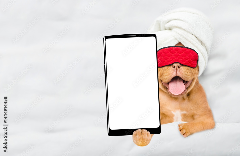 Fototapete yawning mastiff puppy wearing towel and sleeping mask lying on a bed at home and showing big smartph