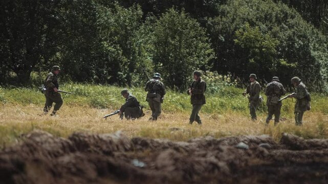 World War II battle reenactment. German Waffen-SS troops retreat and fire back, infantry move across open field near forest. Defense. Offensive of Allied forces or Red Army. Summer military show event