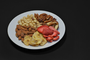 A variety of nuts and dried fruits on a plate on black background. Hazelnuts, pecans, brazil nuts, almonds, dried strawberries and bananas. The concept of nutrition, diet, healthy eating and sports