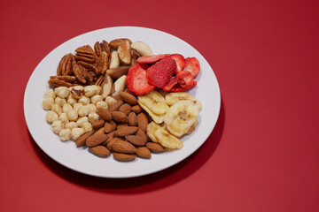 A variety of nuts and dried fruits on a plate on red background. Hazelnuts, pecans, brazil nuts, almonds, dried strawberries and bananas. The concept of nutrition, diet, healthy eating and sports