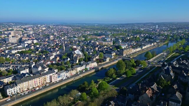 Lateral aerial shot over Le Mans showing the Sarthe river, a bridge with traffic, urban buildings, greenery, and a blue sky.