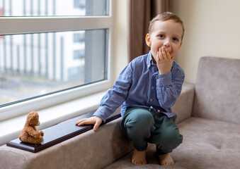 Surprised little boy sitting by window indoors. Young child covering mouth in a surprised gesture...