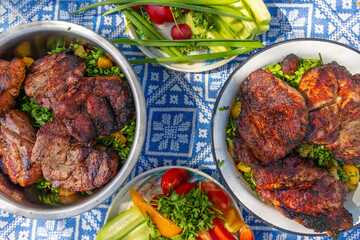 Grilled meat with fresh vegetables on outdoor table. Top view of grilled pork steaks served with herbs, roasted potatoes, and fresh garden vegetables on a patterned outdoor tablecloth.