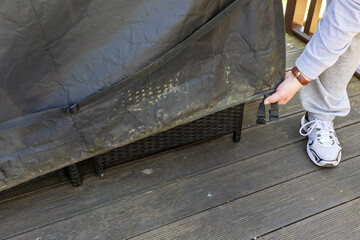 A man lowers a black winter cover onto a patio table on a wooden deck. A hanging strap and buckle...