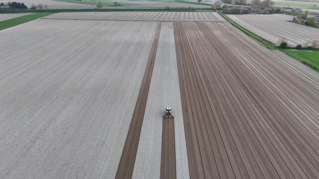 Drone captures a tractor leveling vast fields in Olza, Castelvetro, PC, creating broad parallel bands of light and dark soil that stretch toward the horizon, forming a geometric agricultural pattern.