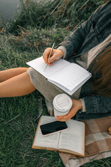 Obraz premium Woman sitting on blanket in grass, writing in a journal with coffee cup in hand. Cozy autumn mood with boots, book, smartphone, and fallen leaves around her