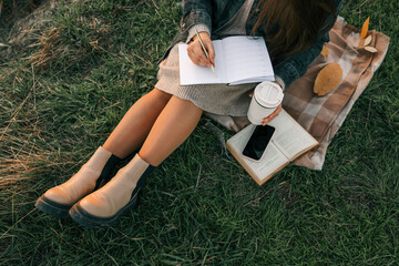 Woman sitting on blanket in grass, writing in a journal with coffee cup in hand. Cozy autumn mood...