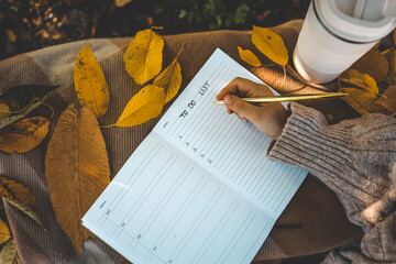 Hand writing in a planner with a gold pen, surrounded by yellow autumn leaves, a reusable cup, and...