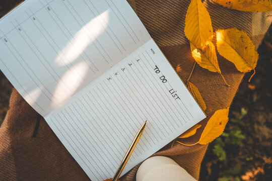Open planner with handwritten "To Do List," gold pen, and scattered autumn leaves on cozy blanket, with warm natural lighting and soft focus background