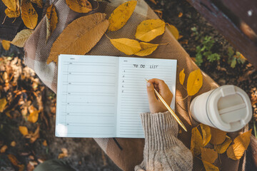 Hand writing in a planner with a gold pen, surrounded by yellow autumn leaves, a reusable cup, and cozy textures, creating a warm seasonal to-do list scene