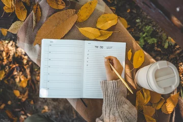 Fotobehang Herfst Hand writing in a planner with a gold pen, surrounded by yellow autumn leaves, a reusable cup, and cozy textures, creating a warm seasonal to-do list scene  © shine.graphics