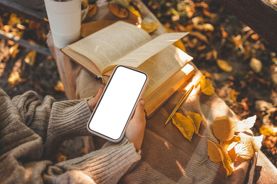 Person in cozy sweater holding smartphone with blank screen next to open book, golden pen, and autumn leaves on blanket in soft natural light