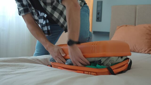 Man trying to close over packed orange suitcase before trip. Male traveler has trouble while packing a lot of clothes, preparing for travel