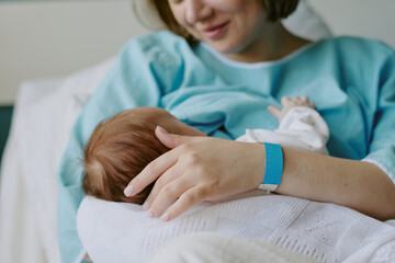 New mother in hospital gown breastfeeding newborn baby while holding closely. Both mother and baby...