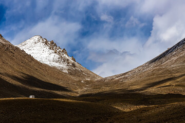 A wide-angle view of a small white cabin amidst shadows of clouds at the foothills of Wari La mountain pass in Ladakh, India.