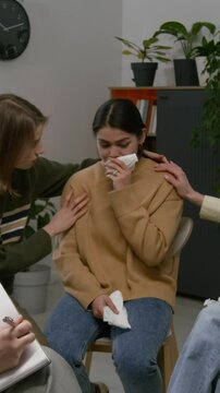 Vertical handheld zoom out shot of distressed woman crying and receiving comfort at supportive group therapy session