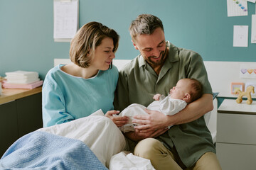 Obraz premium Caucasian parents smiling while holding their newborn baby in a hospital room. The mother sits on the bed covered with a light blanket