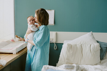 Mother in hospital gown holding newborn baby while standing by window in hospital room. Background...