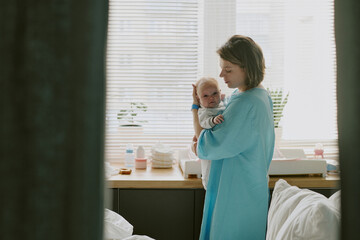 Mother standing near window holding her newborn baby in recovery room without smiling. Toddler...