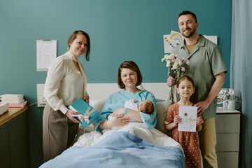 Happy family celebrating newborn arrival together in hospital room, posing with presents and...