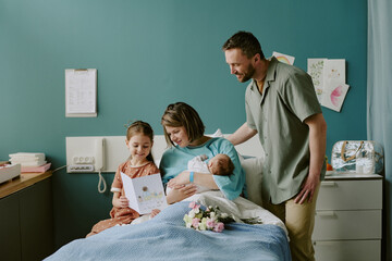 Happy family welcoming new baby in hospital room with mother holding newborn, father and daughter...