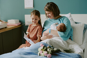 Mother sitting in hospital bed, holding newborn, while daughter sits nearby with a card. Room...