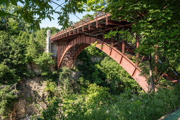 Scenic red steel arch bridge spanning a lush green ravine, surrounded by dense forest. A perfect example of engineering blending into nature, photographed on a bright summer day.
