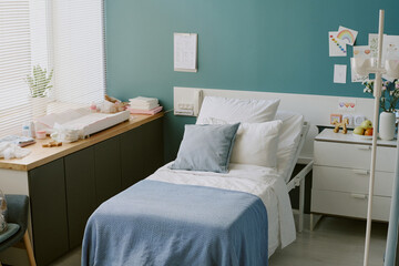 Comfortable bedroom with pastel decor, neatly made bed with blue blanket, and various decorations adorning walls and bedside table. Soft natural light entering through blinds
