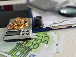 A digital scale shows several gold rings and chains alongside 100 euro banknotes arranged neatly