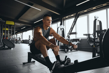 Determined attractive male using rowing machine in contemporary gym. Fit young man working out on...