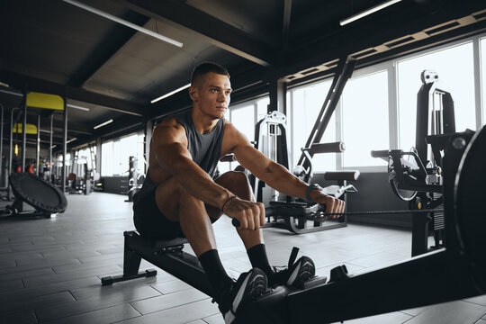 Determined attractive male using rowing machine in contemporary gym. Fit young man working out on rowing equipment at fitness center. Weight loss and cardio exercise concept - Powered by Adobe